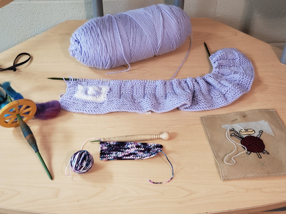 A drop spindle, a crocheted swatch, a half-finished needlepoint, and an in-progress baby blanket