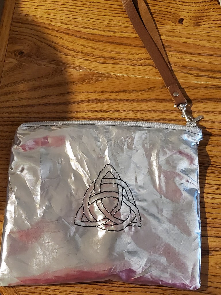 Silver clutch with a Celtic knot embroidered on it. It has a brown leather zipper pull.