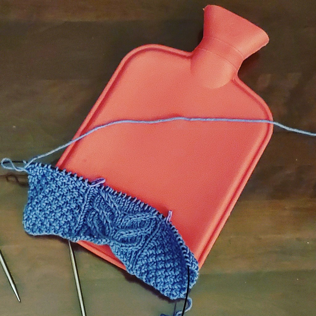 A hot water bottle, covered by a bit of purple, cable knitting on needles